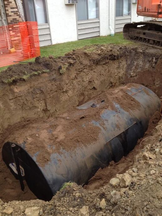 A buried, cylindrical oil tank unearthed in a trench, next to a building and construction equipment.