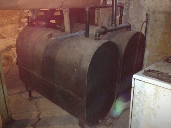 Two dark oil tanks in a basement. Pipes connect to the tanks. White appliance visible on the right.