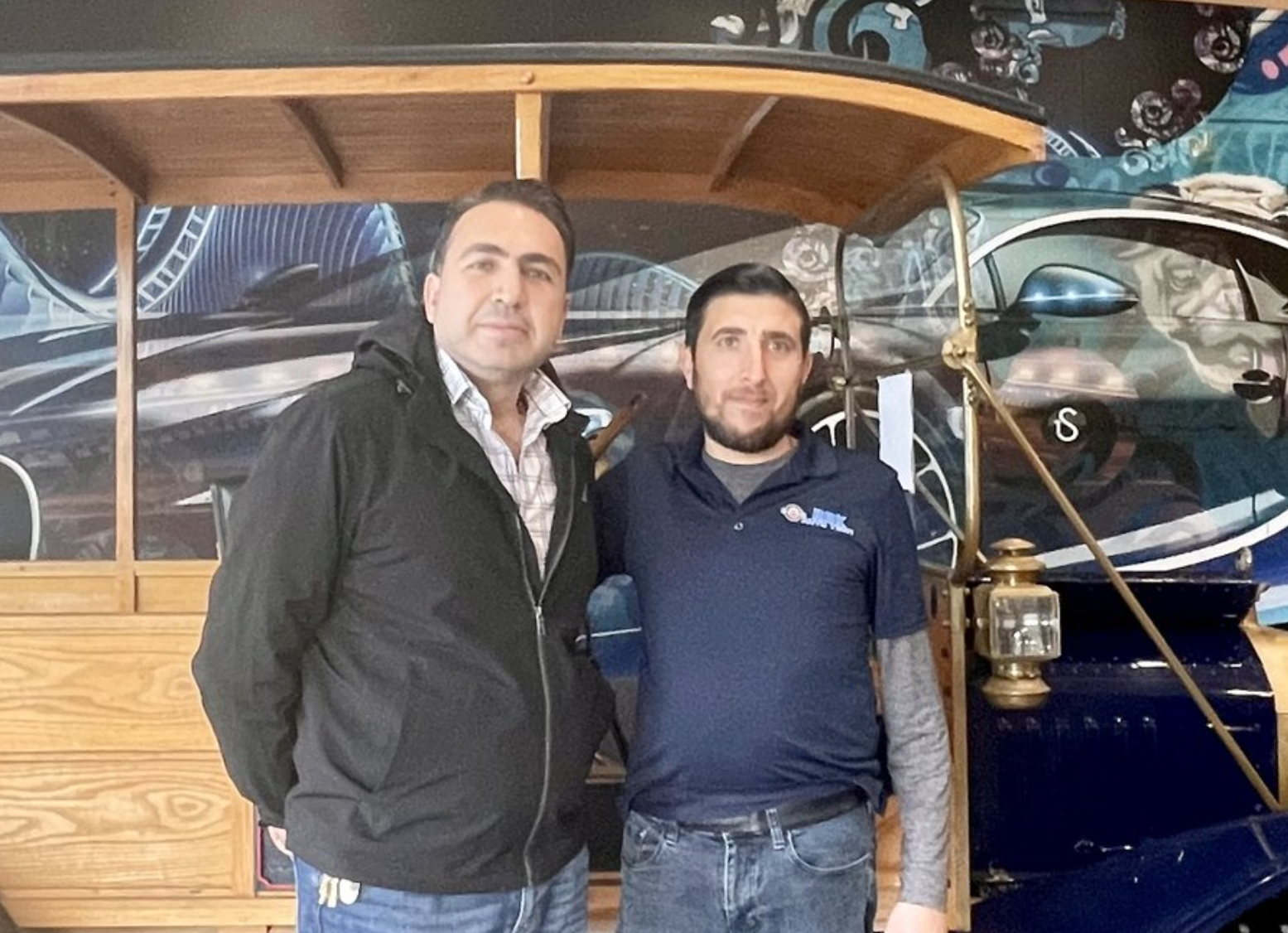 Two men posing beside a vintage blue car. One wears a black jacket, the other a navy shirt. Behind them, a decorated wall.
