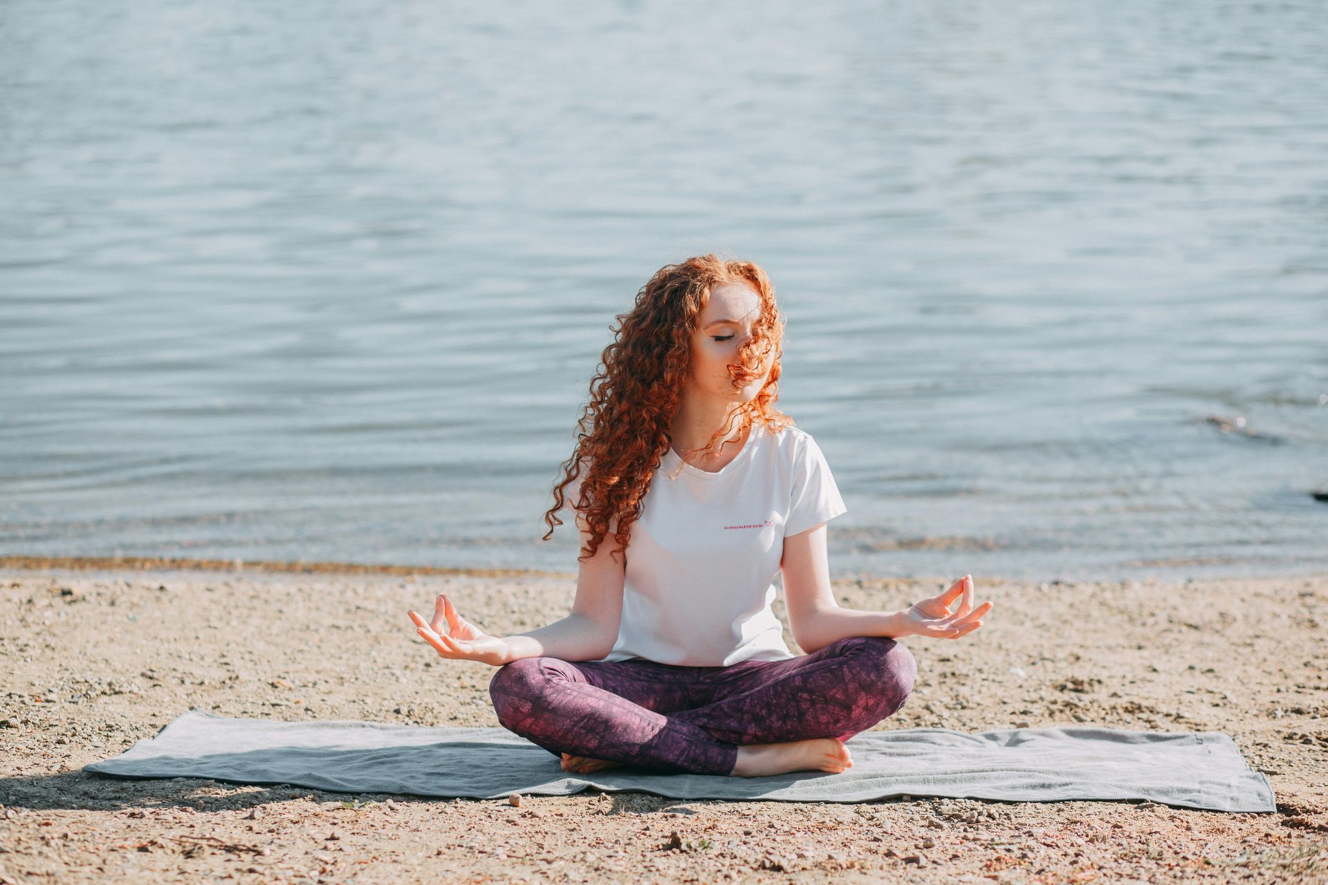 A woman is sitting in a lotus position on a yoga mat on the beach.