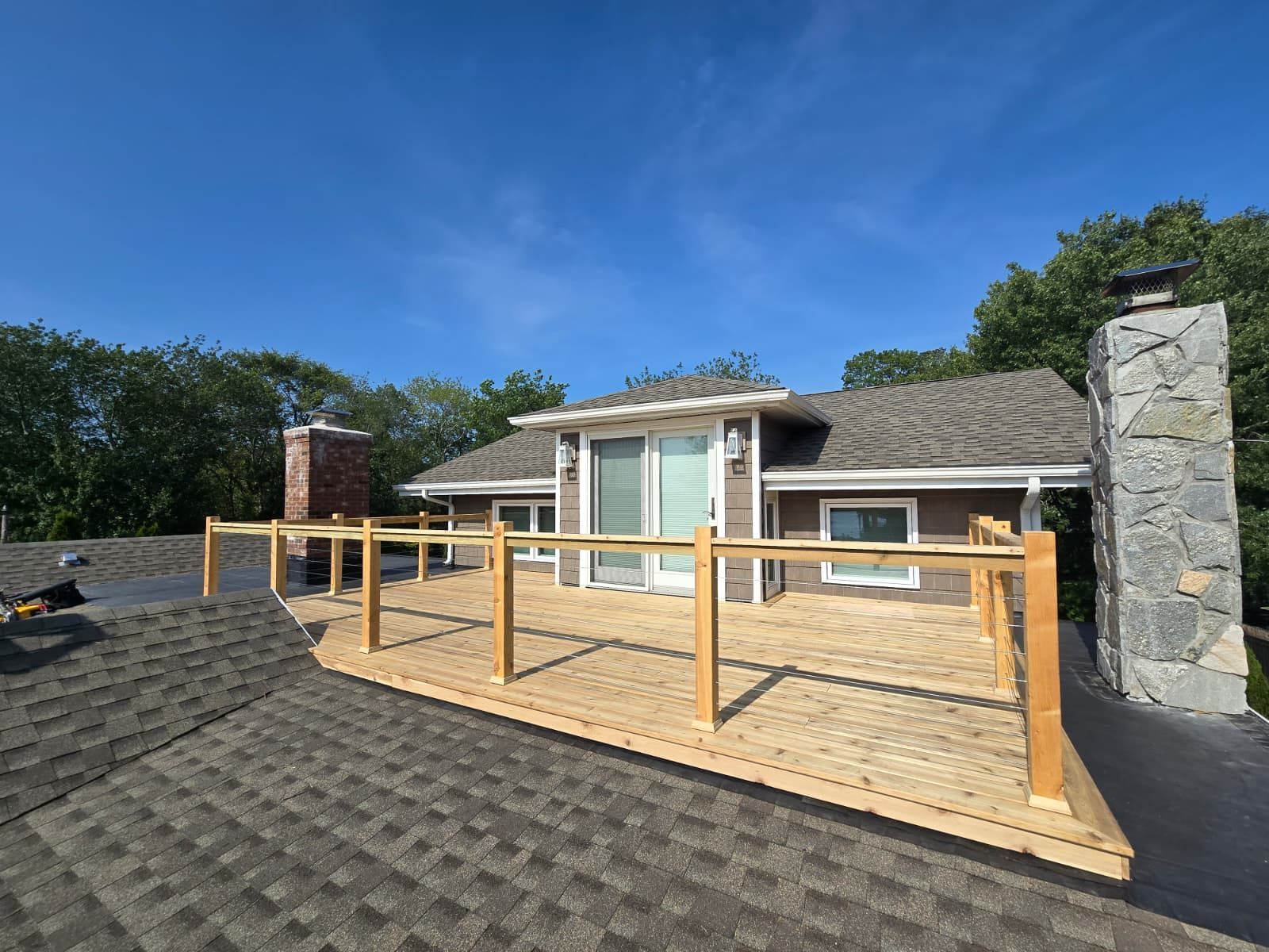 A wooden deck with a railing on a rooftop with a house, chimney, and blue sky.