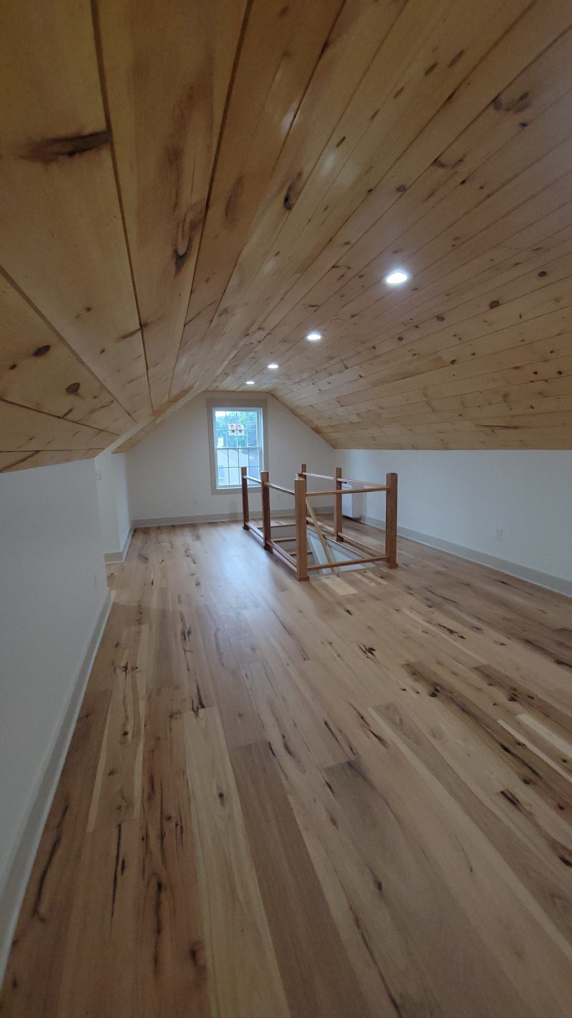 Loft with wooden floor and ceiling, white walls, small window, and light-colored wooden furniture.