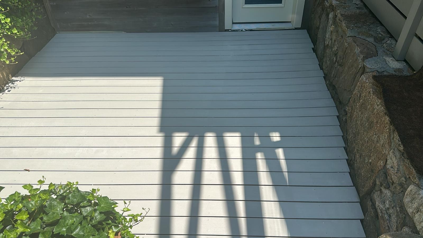 Gray painted wooden deck in front of a white door, with shadows and stone wall on the side, greenery at the bottom.