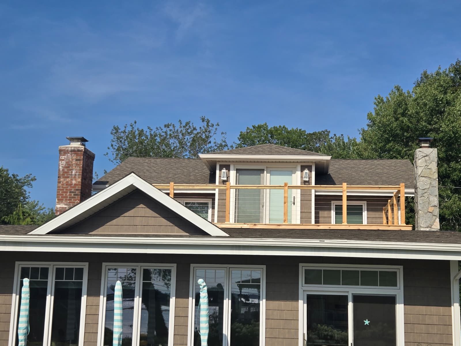 Two-story house with a deck on the roof, featuring brown siding and windows, against a blue sky.