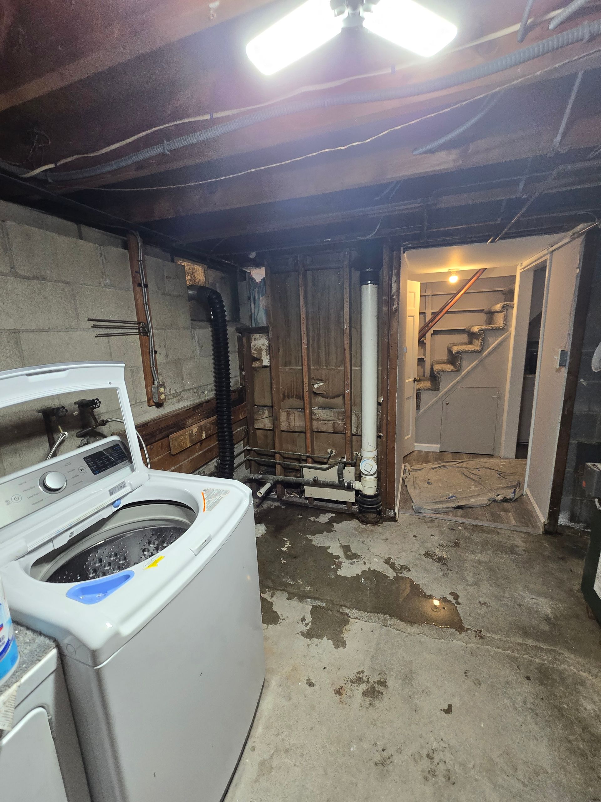A wet, unfinished basement with a washing machine, pipes, and stairs. Water damage visible.