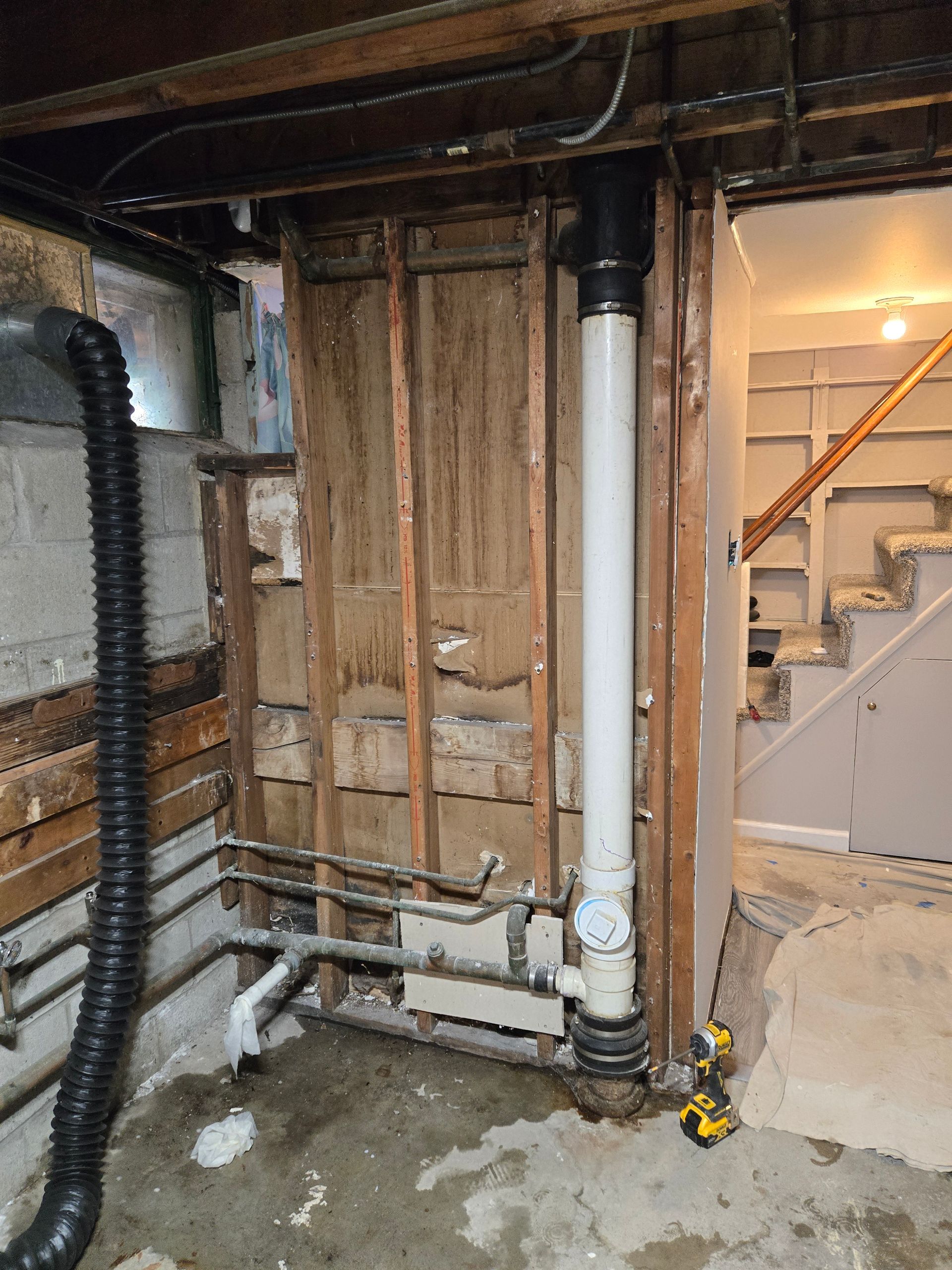 Basement renovation: Exposed plumbing and studs, white pipe, black vent, stairs in background.