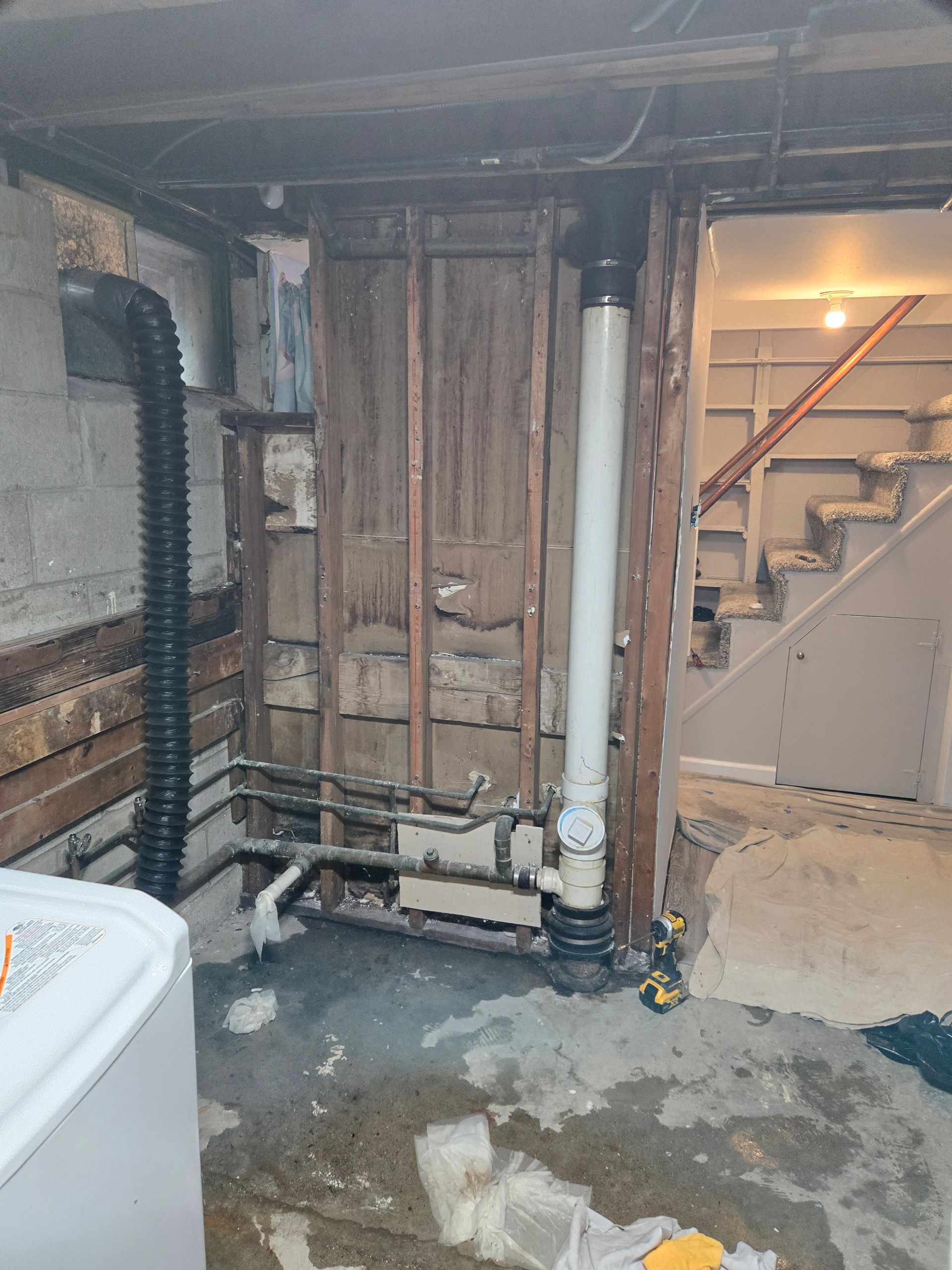 Basement undergoing renovation. Exposed pipes and framing, concrete floor, washing machine, and stairs in the background.