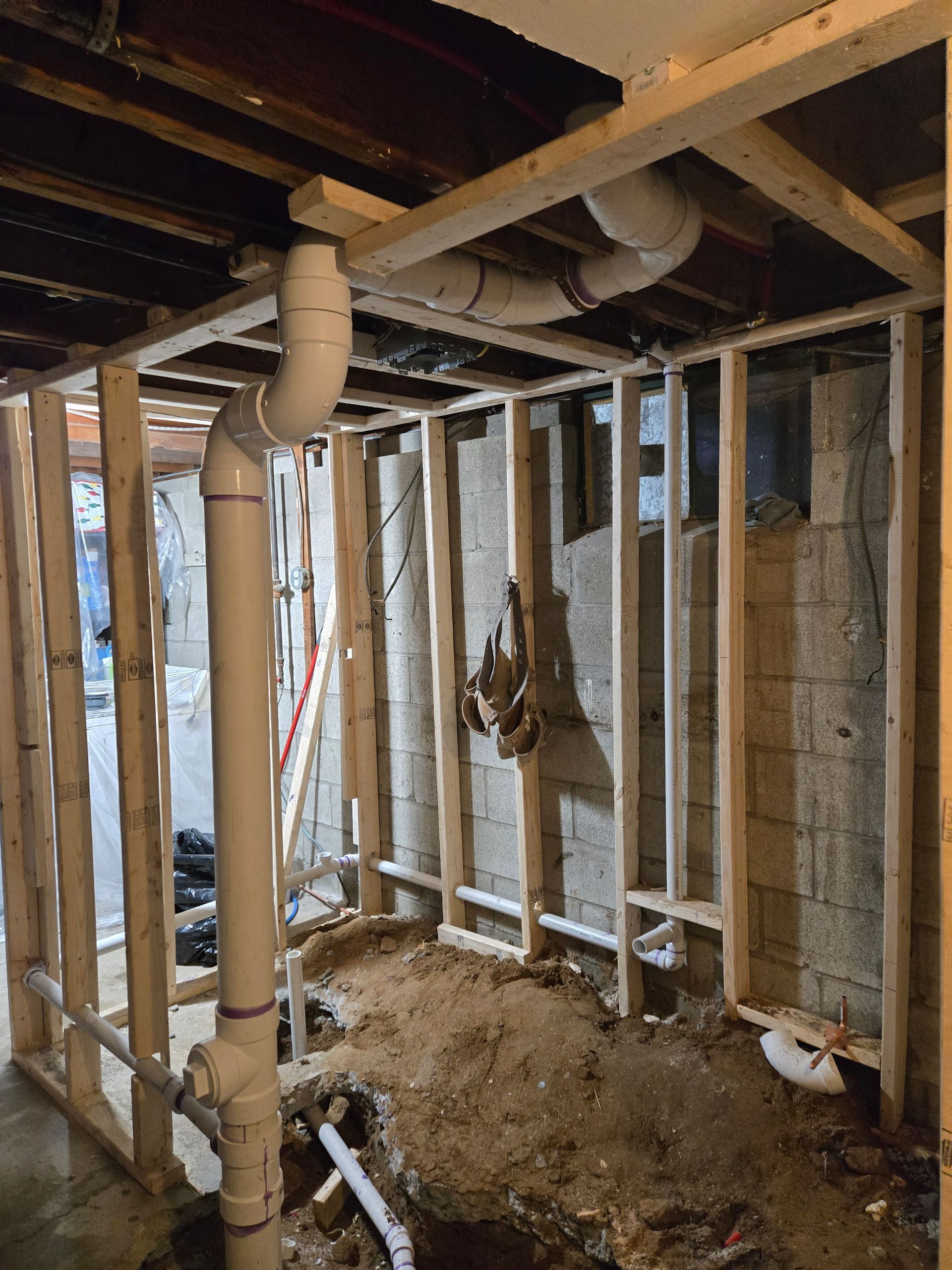 Basement construction with exposed plumbing and wooden framing.  Concrete wall in background, dirt on the floor.