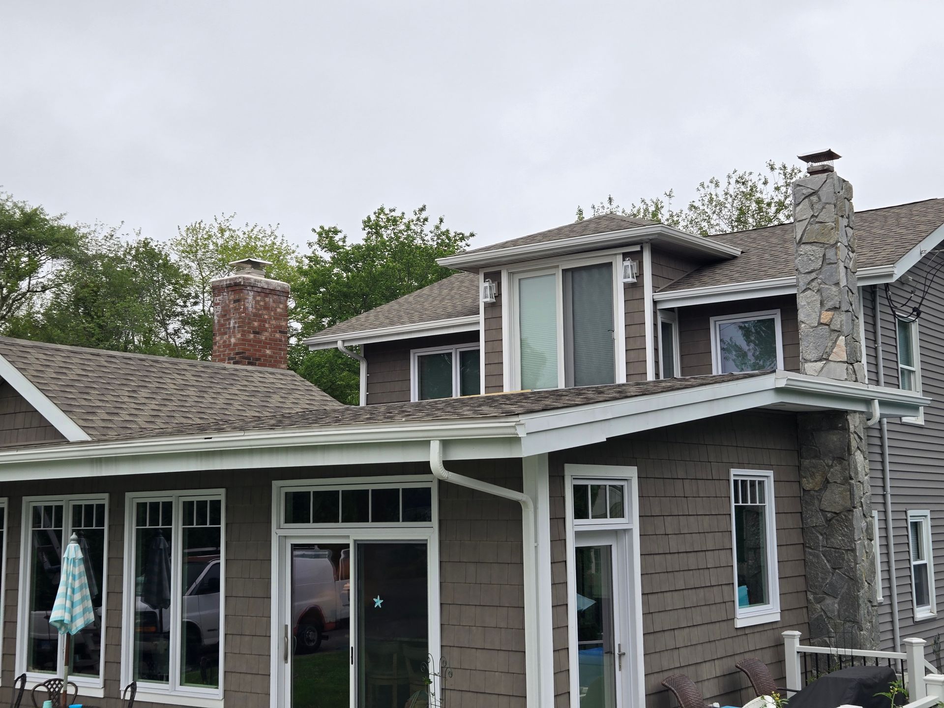 Brown shingled house with white trim, windows, and stone chimney against a cloudy sky.