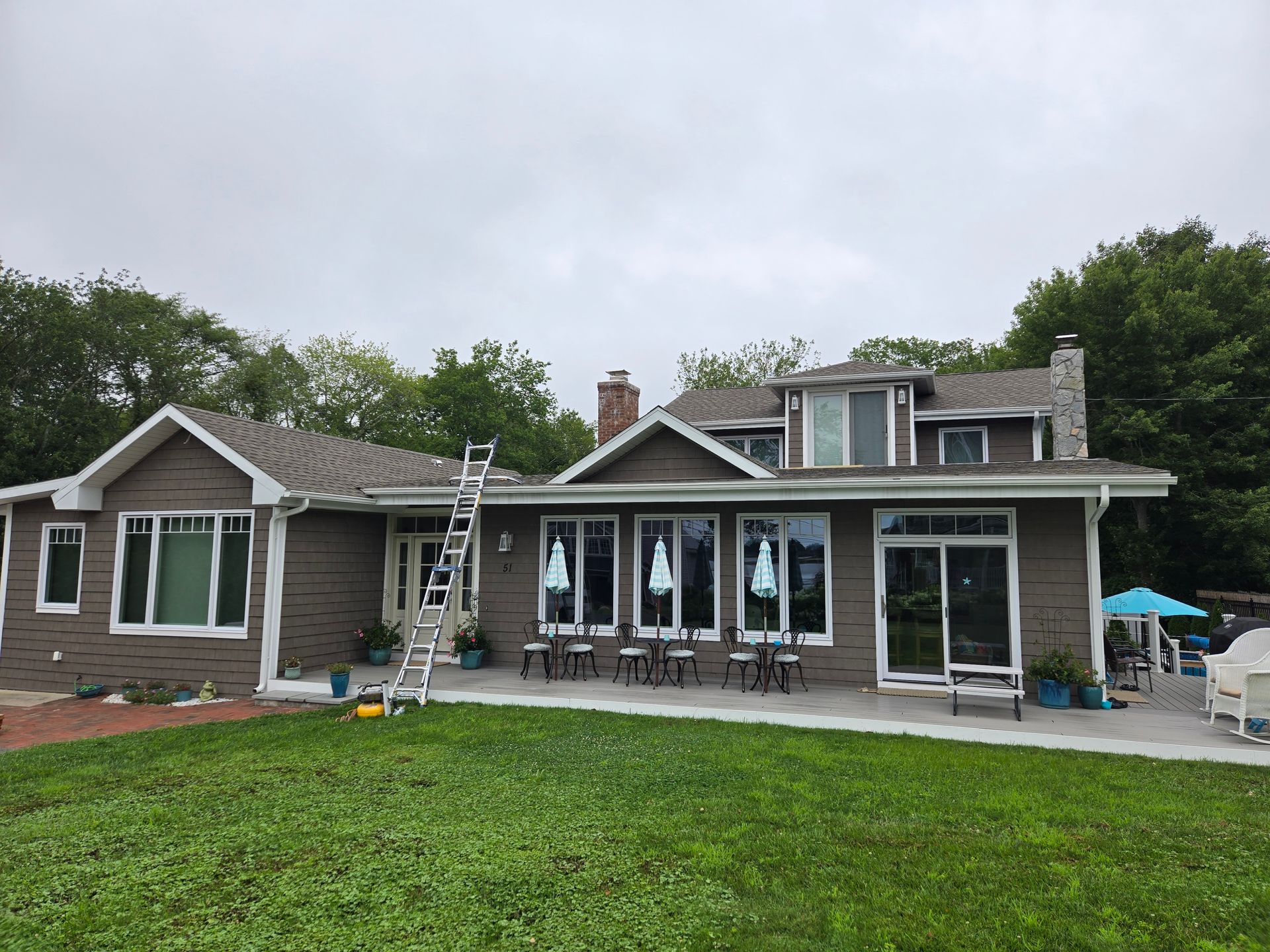 Brown house with white trim, gray roof, and ladder leaning against the side, on a cloudy day.