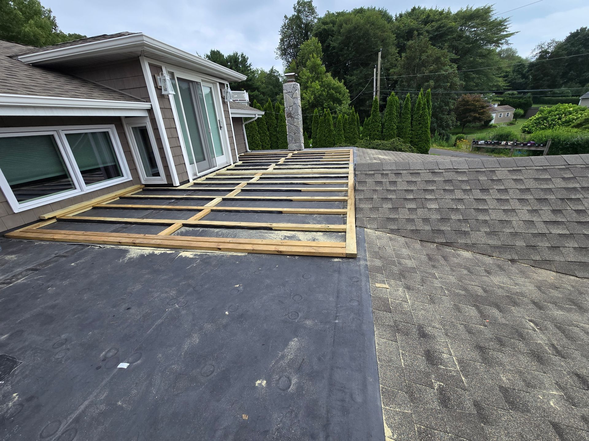 Roof with exposed wooden deck frame being repaired; gray asphalt shingles, green foliage in background.