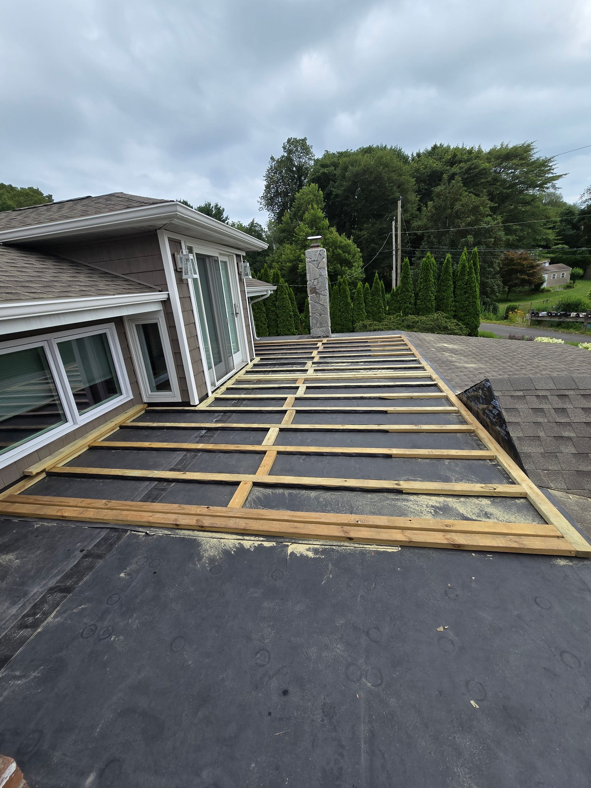 Rooftop with wooden framework under construction. Building sits near a chimney and trees. Gray sky overhead.