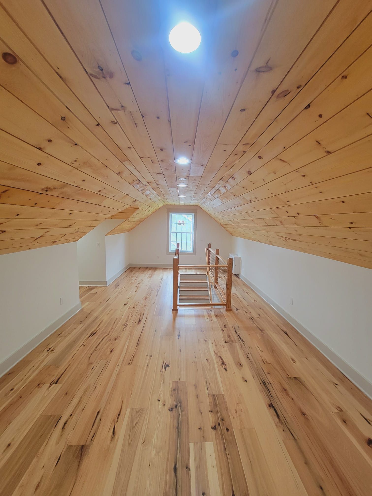 Wooden floored attic room with sloped wood ceiling and white walls, window in far wall.