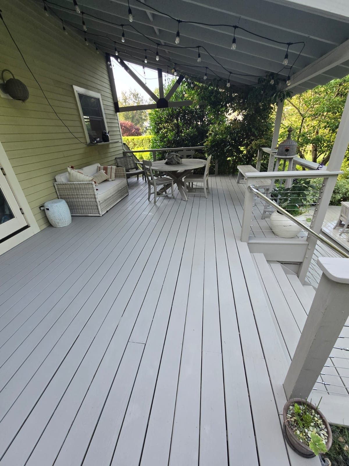 A gray wooden deck with outdoor furniture, overhead string lights, and a lush green backdrop.