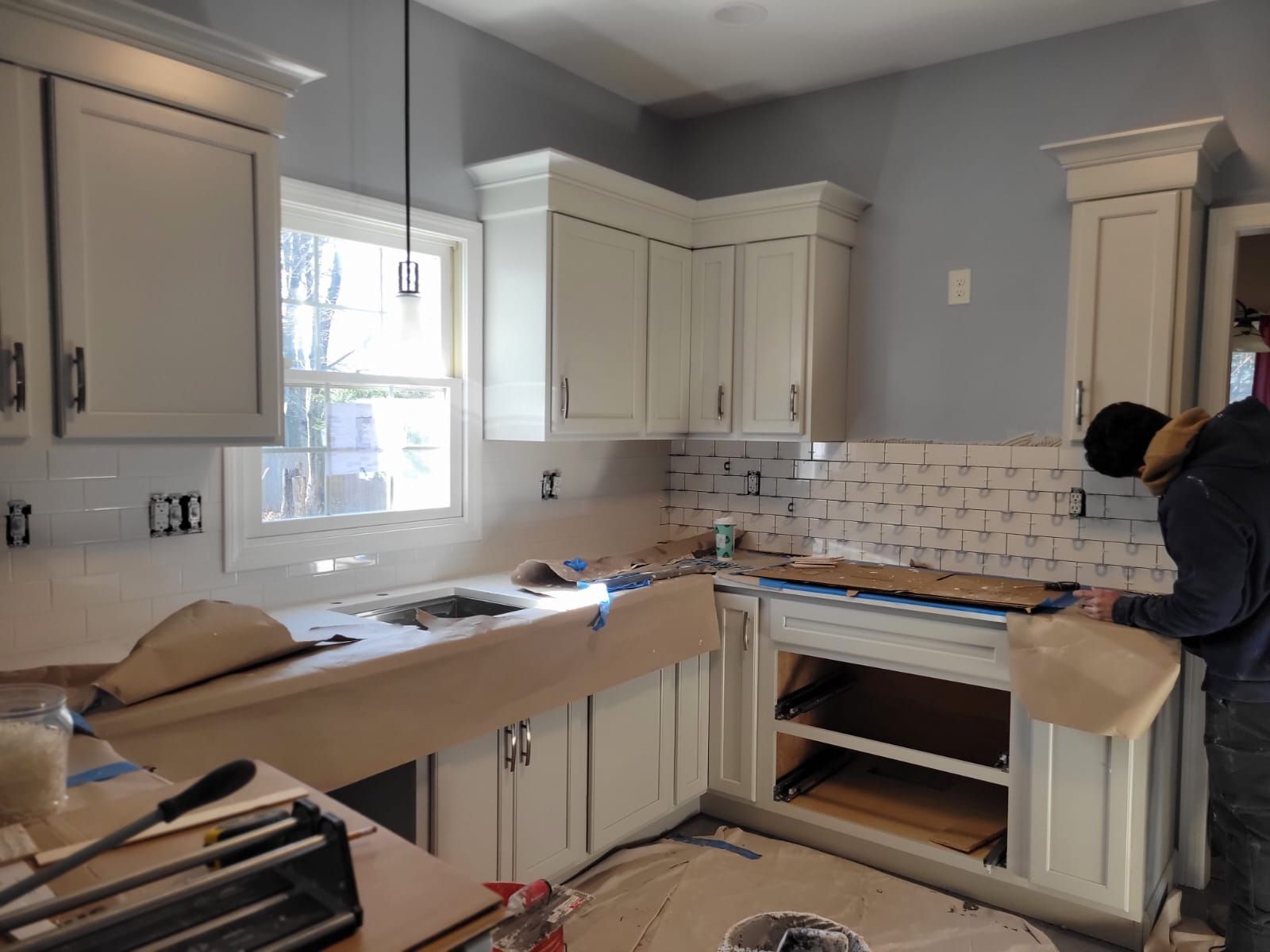 Kitchen renovation: Off-white cabinets, tiling backsplash, worker installing tiles near a window, with light blue wall.