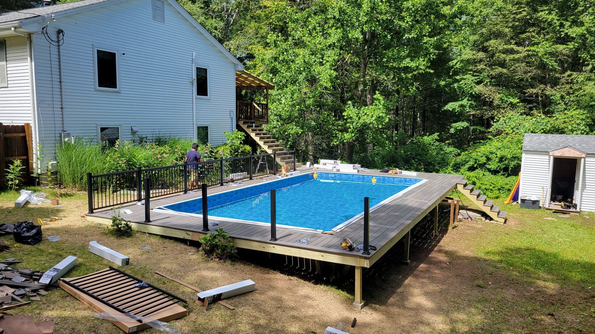 Above-ground pool with dark deck, black railing, and a person near the pool in a backyard setting.