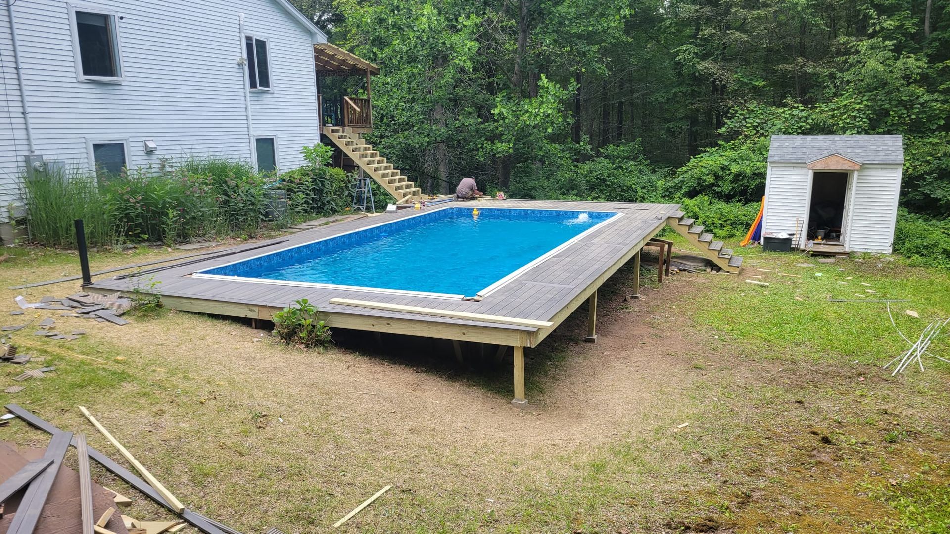 Above-ground pool with blue water, gray deck, and shed in a grassy backyard. A house and stairs are visible.