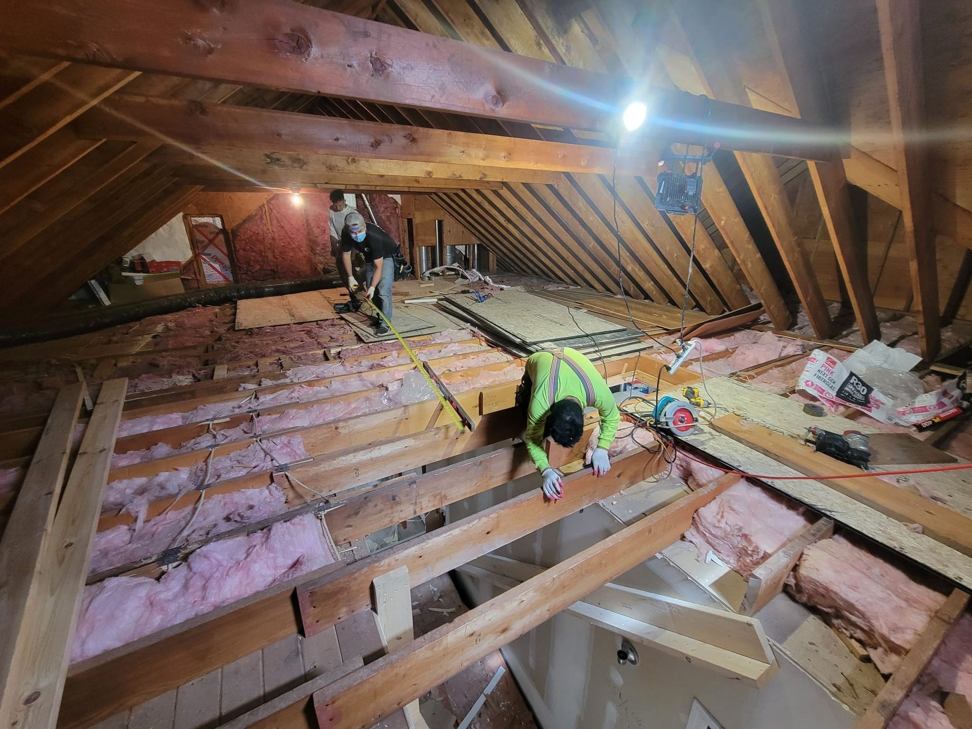 Two workers in an attic removing old insulation and wooden boards. Light illuminates the space.