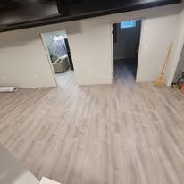 Basement with light wood-look flooring, white walls, and doorways leading to other rooms; a broom is near one door.