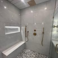 Modern bathroom with white tiles, shower bench, gold fixtures, and pebble floor.