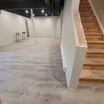 Empty basement with gray flooring and stairs. A white wall with a wooden trim is on the right side.