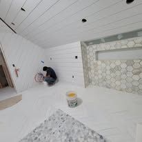 Person installing tile in a bathroom with white plank walls and ceiling. Bucket of grout on the floor.