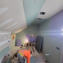 A person in an orange shirt is working on a drywall installation in a room under construction.