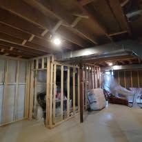 Basement with wooden framing and ductwork; unfinished, with supplies and furniture in the space.