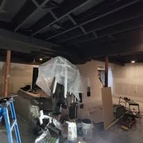 A cluttered basement with exposed beams. A plastic-covered object sits near a ladder, drywall, and unfinished walls.