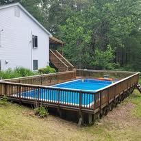 Above-ground pool enclosed by wooden fencing near a house, surrounded by grass and trees.