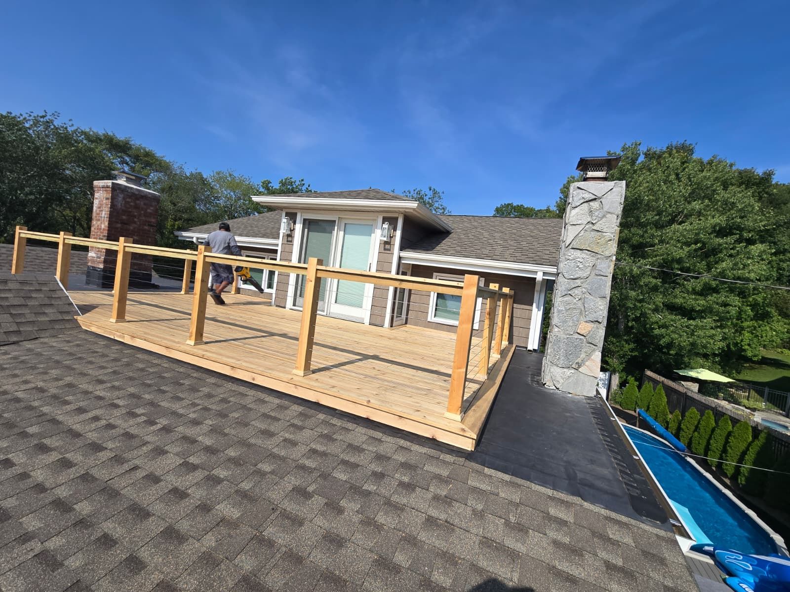 A roof deck with a wood railing is being built. A worker is visible. Trees and pool in view.