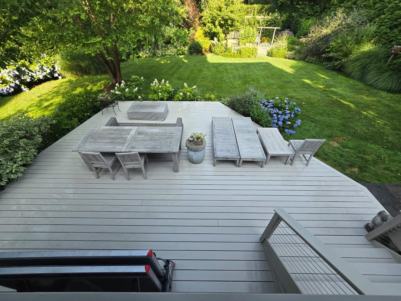 A backyard deck with furniture. Lounge chairs, a dining table, and a fire pit are arranged on the light gray deck.