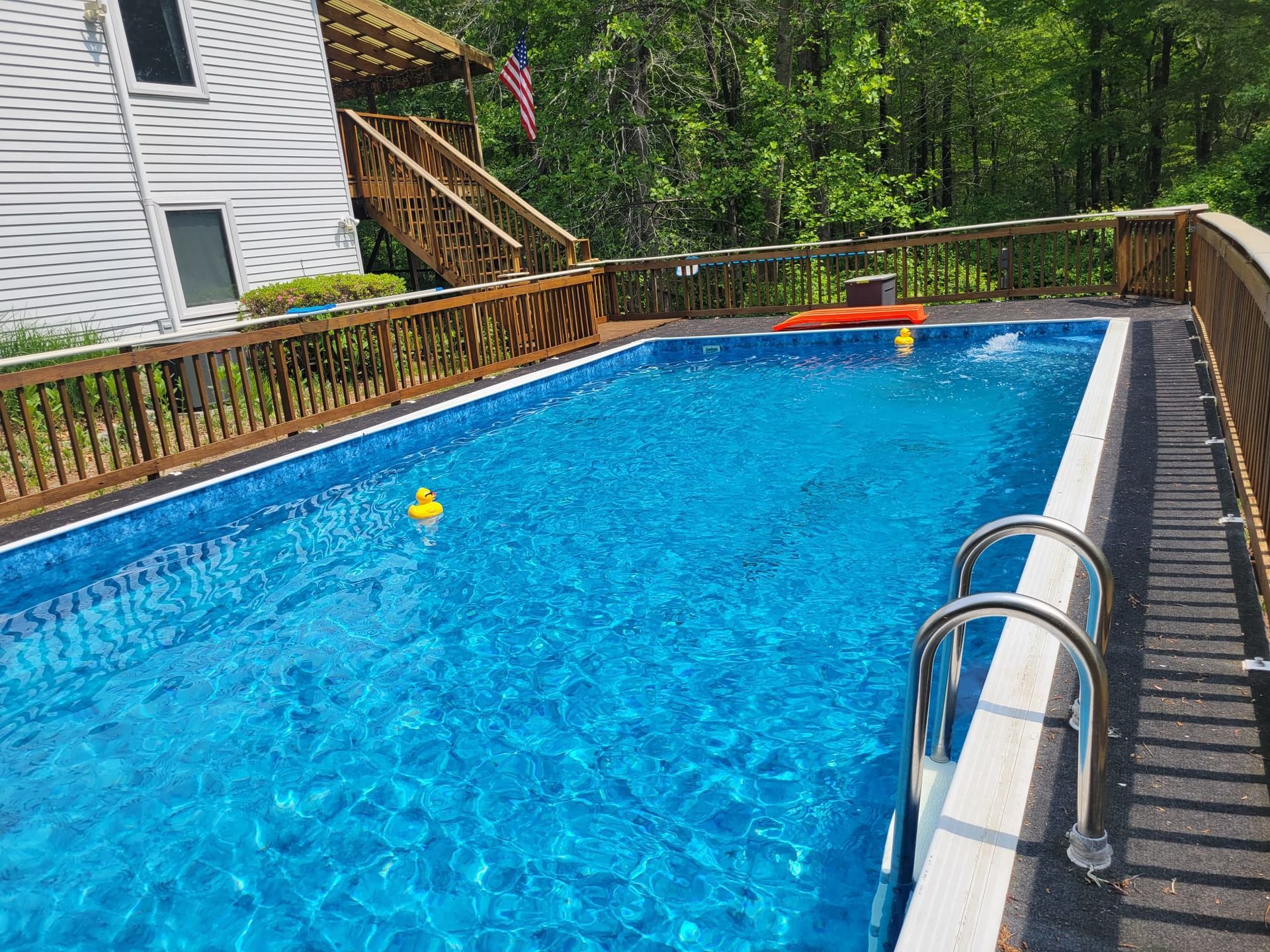 Pool with blue water surrounded by a wooden deck and house, ducks float, sunny day.