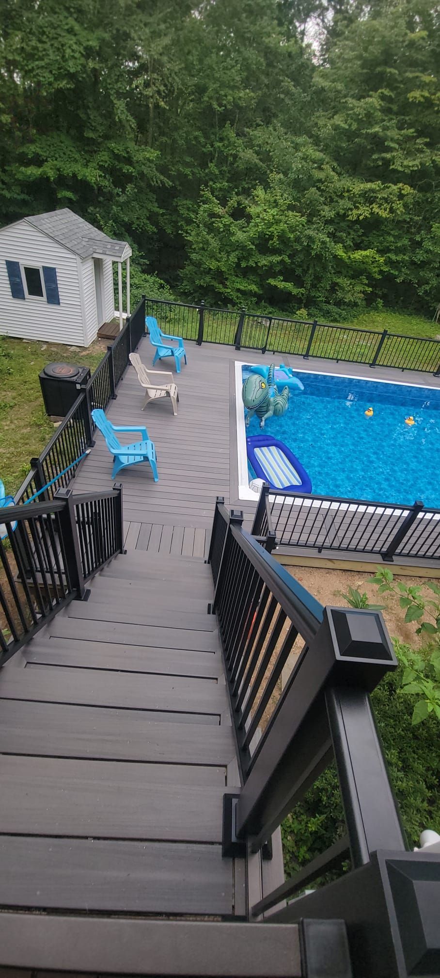 Wooden deck with steps leading down to a pool, surrounded by trees and a small shed.
