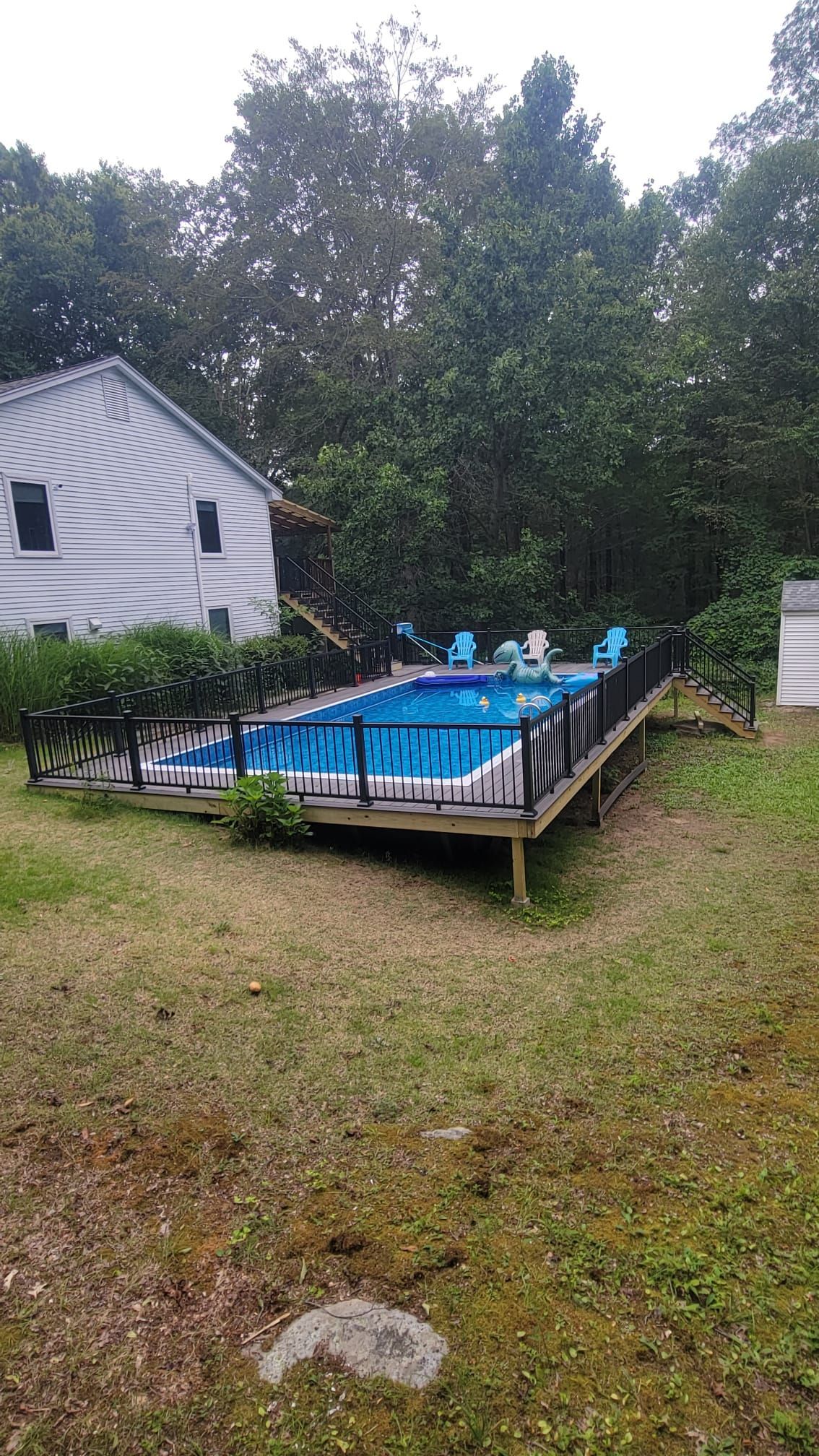 A backyard pool surrounded by a wooden deck, blue chairs, and trees. A white house is to the left.