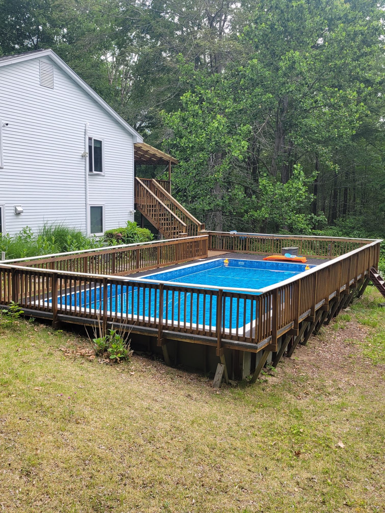 Above-ground pool with wooden deck next to a white house and trees.