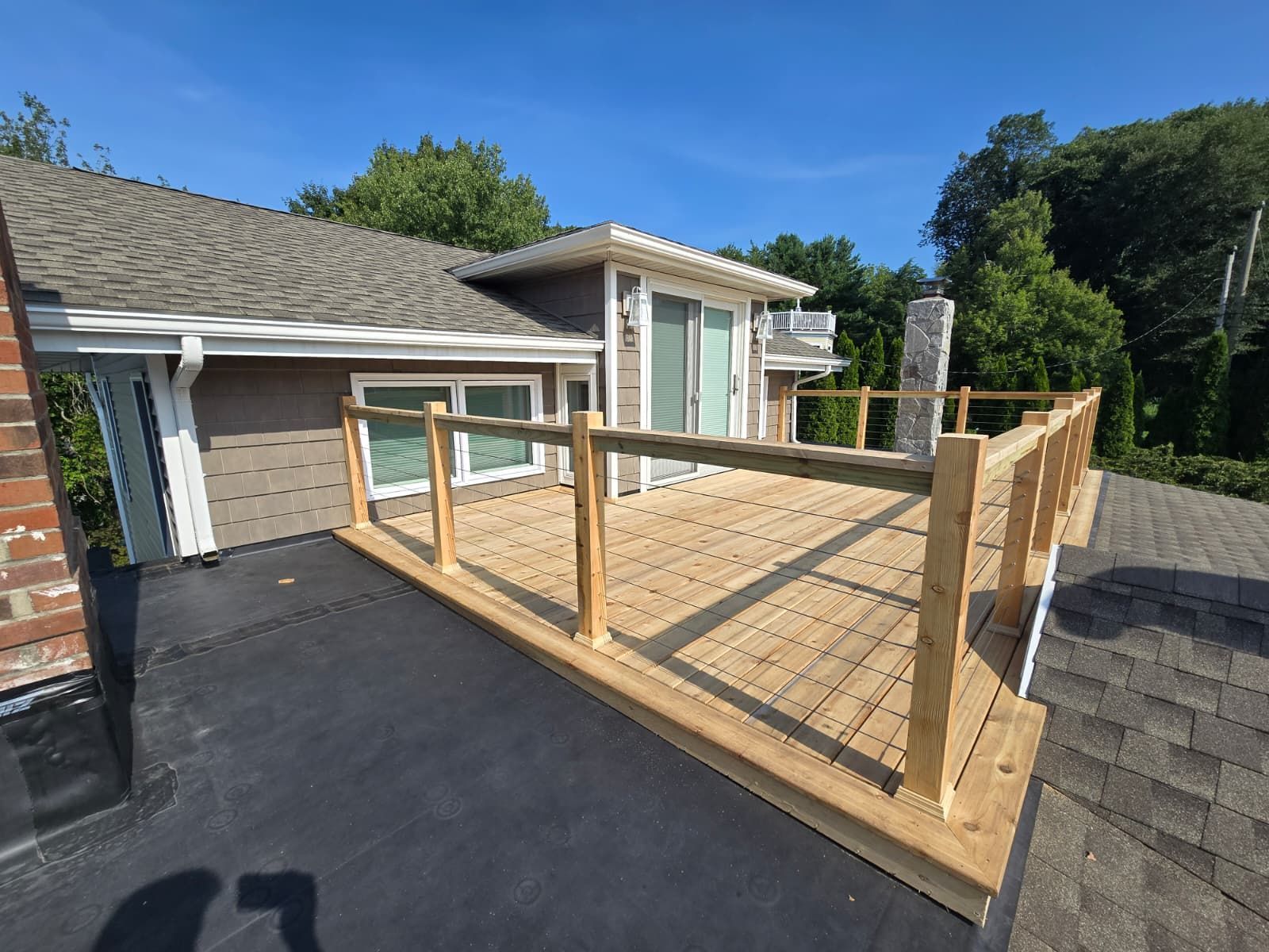 Wooden rooftop deck with railing on a house, surrounded by trees, blue sky.