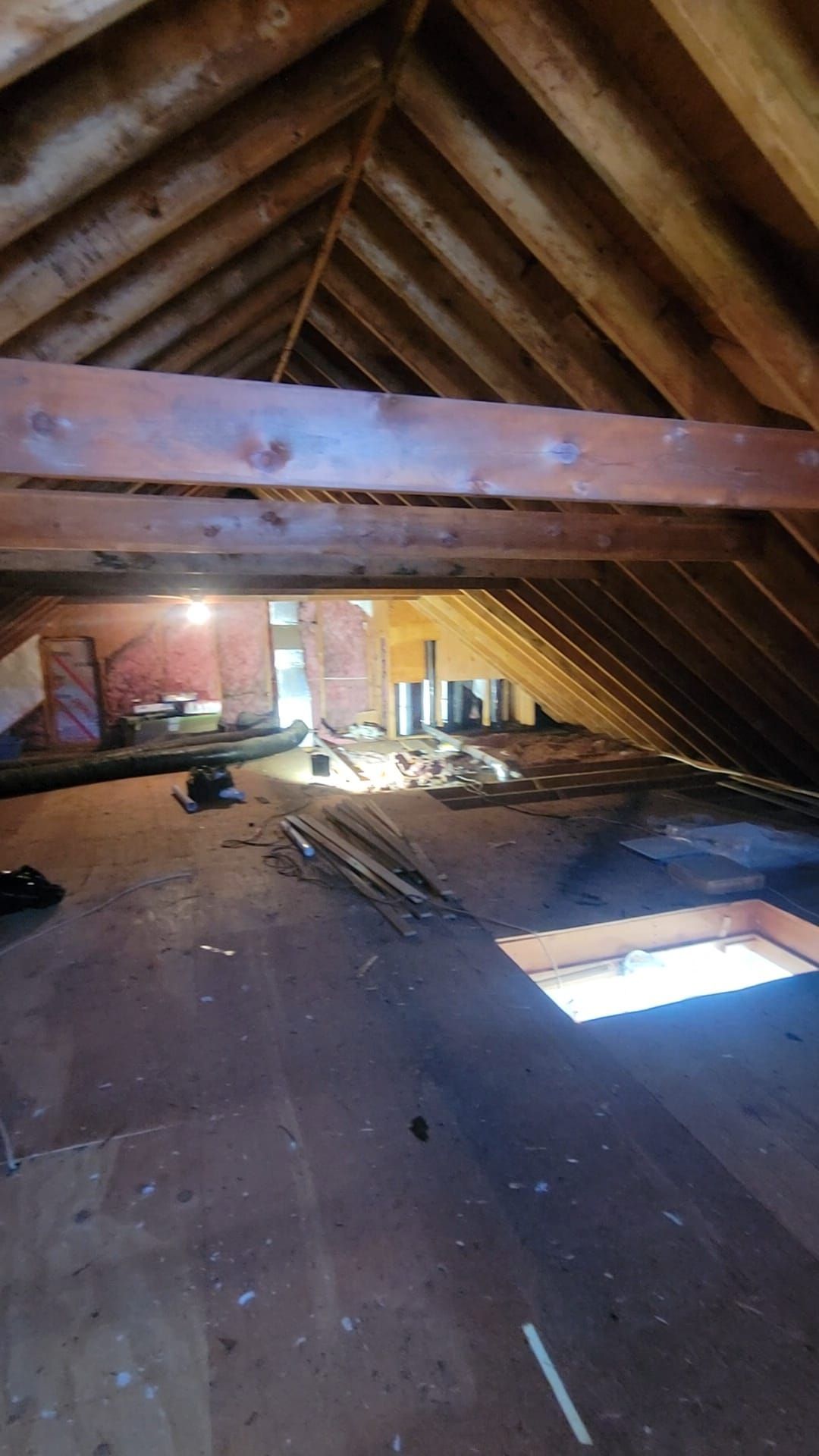 Attic interior with exposed wooden beams, rough flooring, and an open skylight, bathed in natural light.