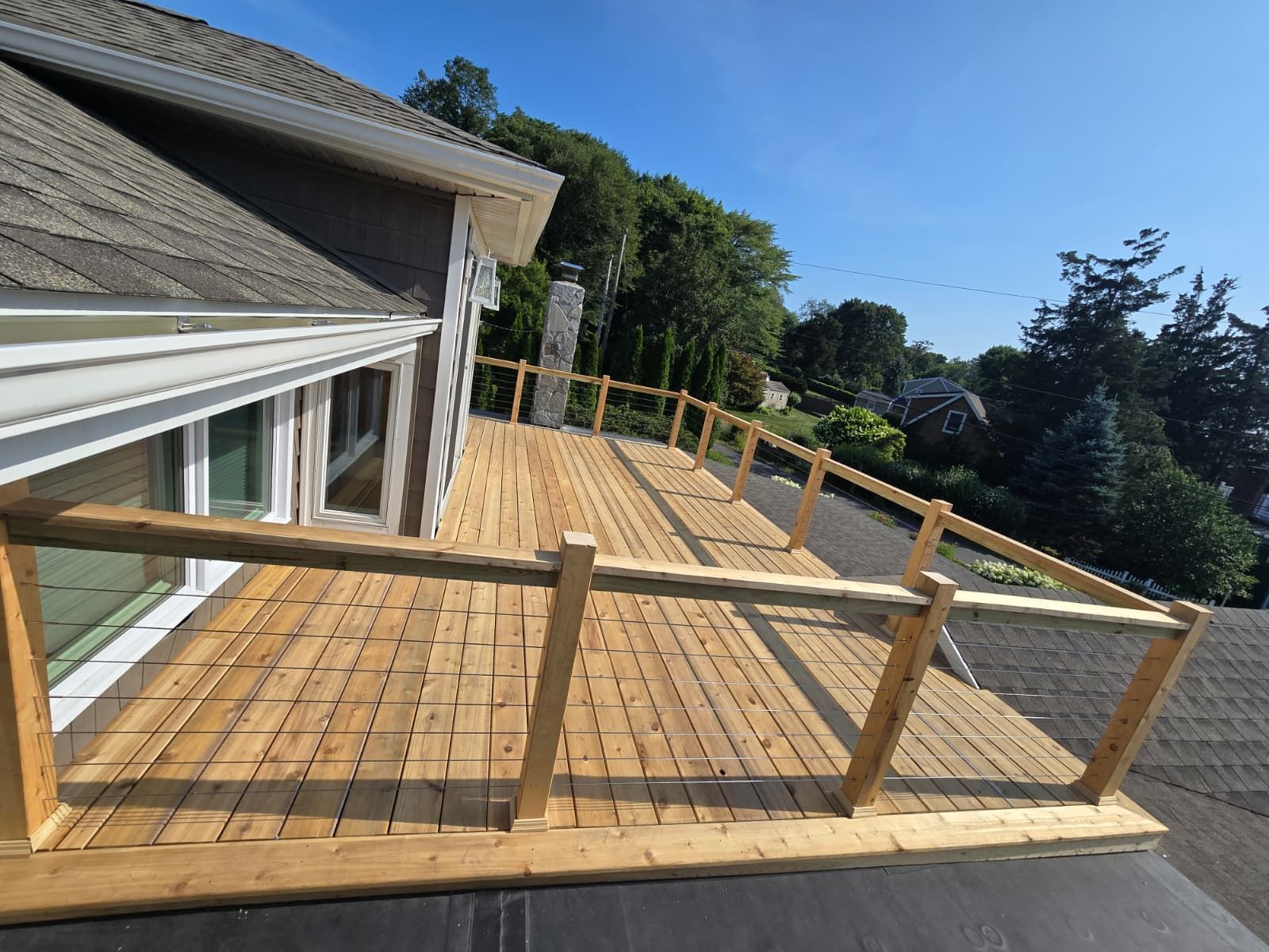 Wooden deck with railing attached to a house, overlooking trees under a blue sky.