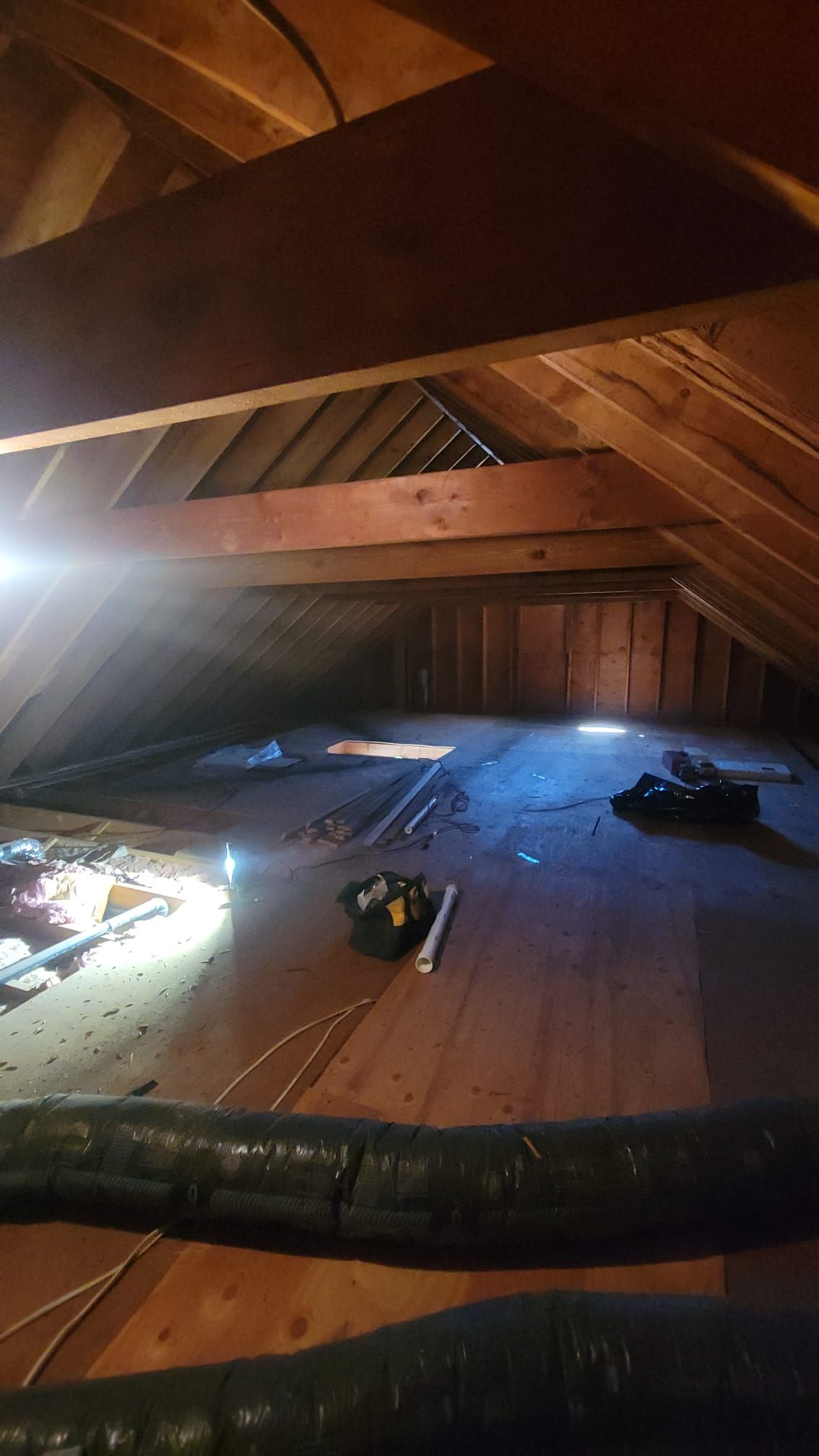 An attic interior with wooden beams, insulation, and some items on the floor; natural light streams in.