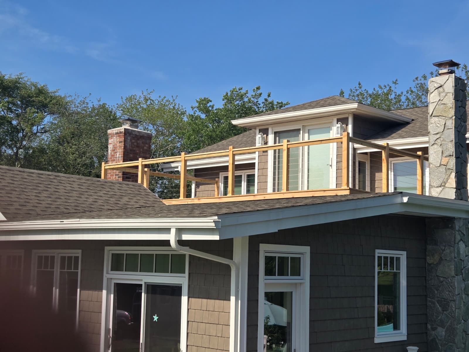Wooden frame construction on a house roof.  The house has a dark gray exterior and a brick chimney. Clear blue sky.