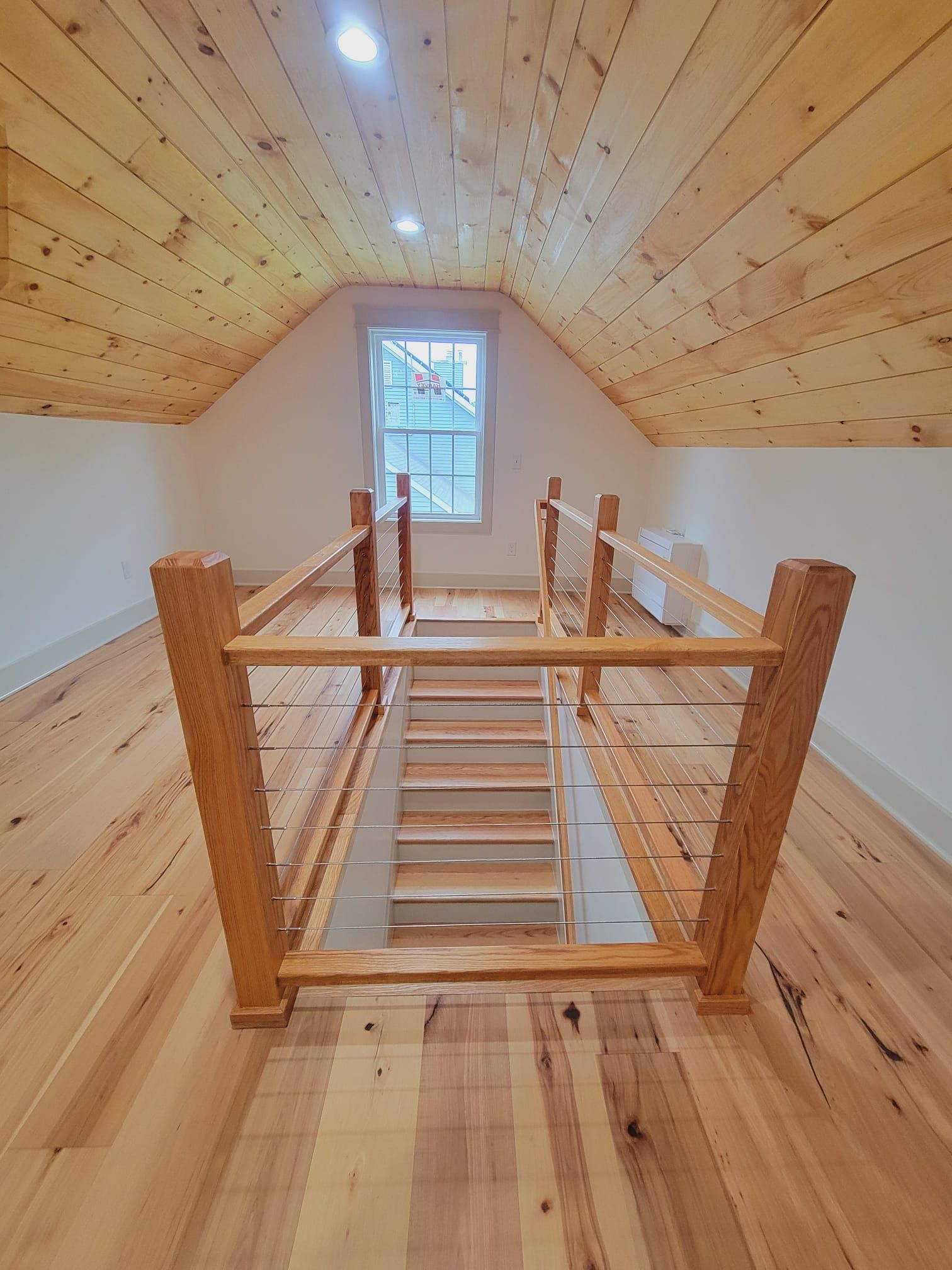 Loft interior with wooden floors and railing, stairs leading down to a lower level, window at the far end.