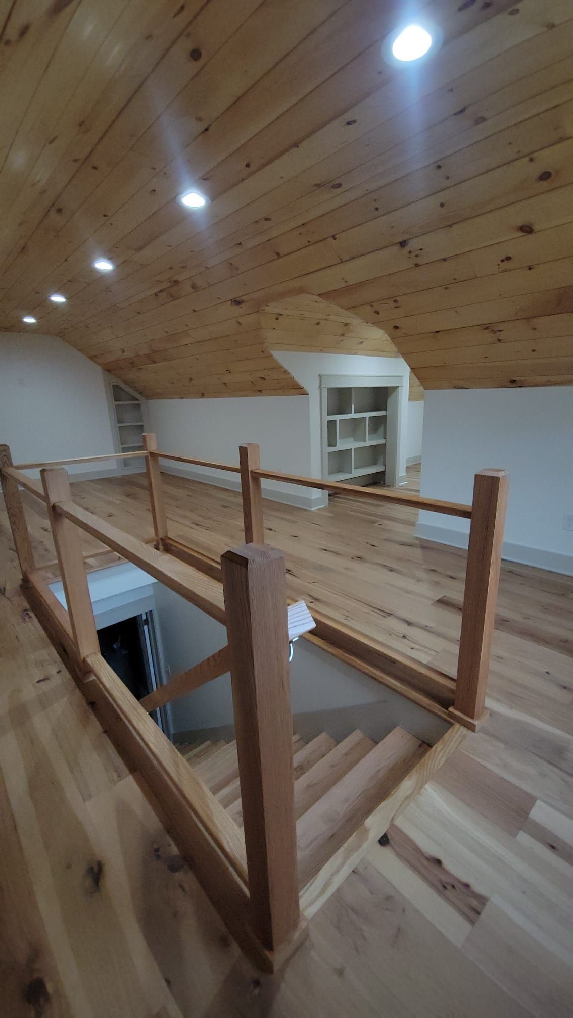 Loft with wooden floor, ceiling, and railing; stairs lead down to a lower level. Shelves in the back.