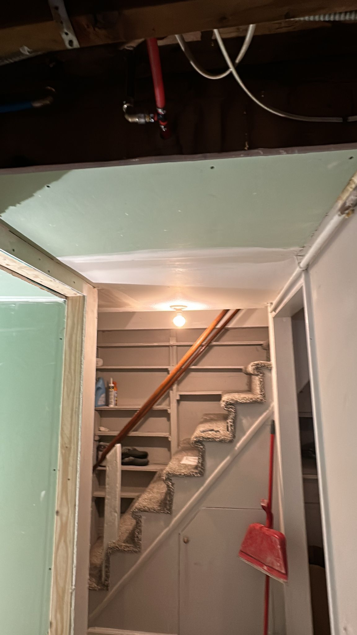 Staircase leading up, viewed from inside a room with unfinished walls and ceiling. A broom hangs on the side.