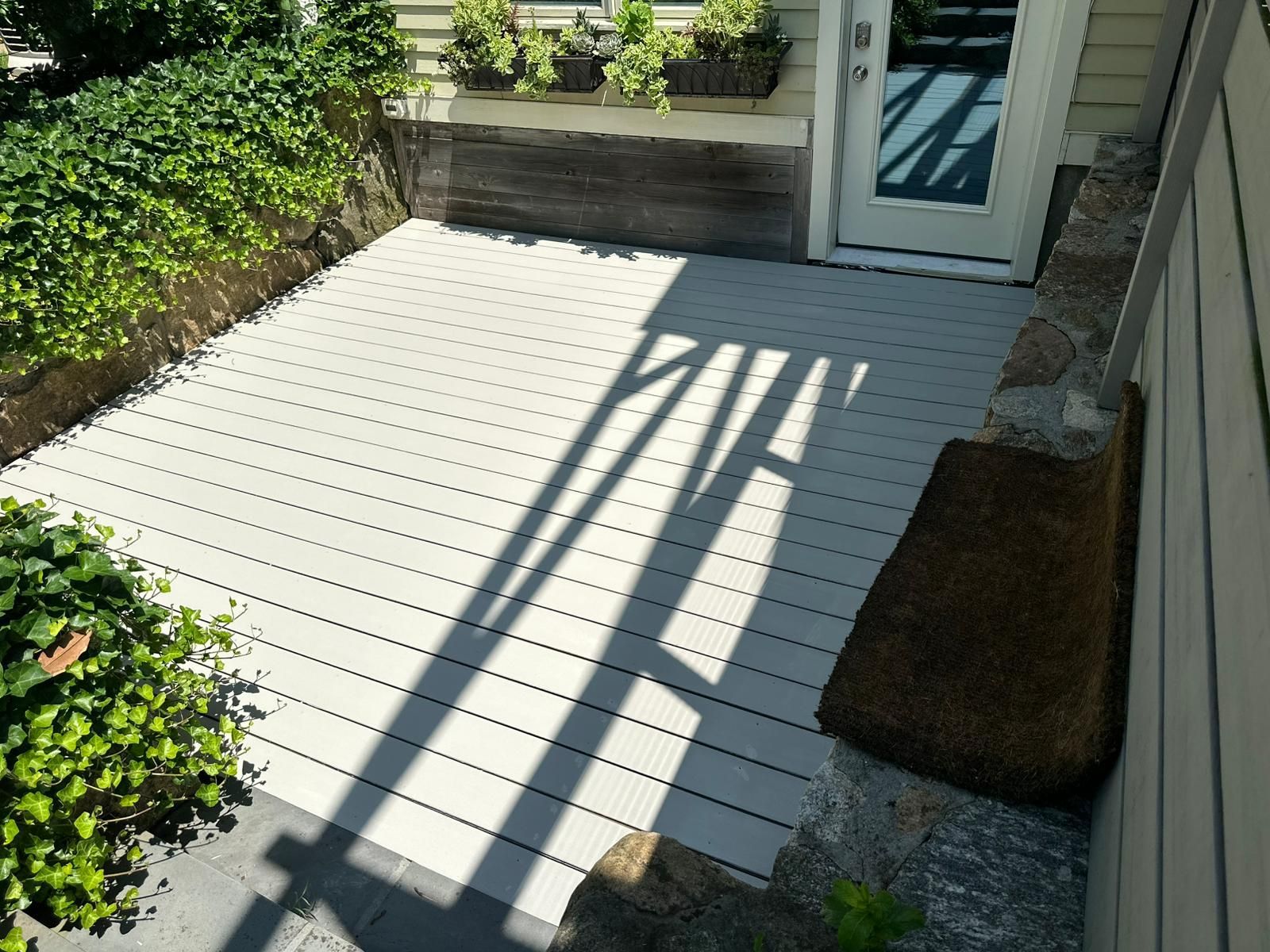 Light gray composite deck next to a door and stone wall, with greenery.