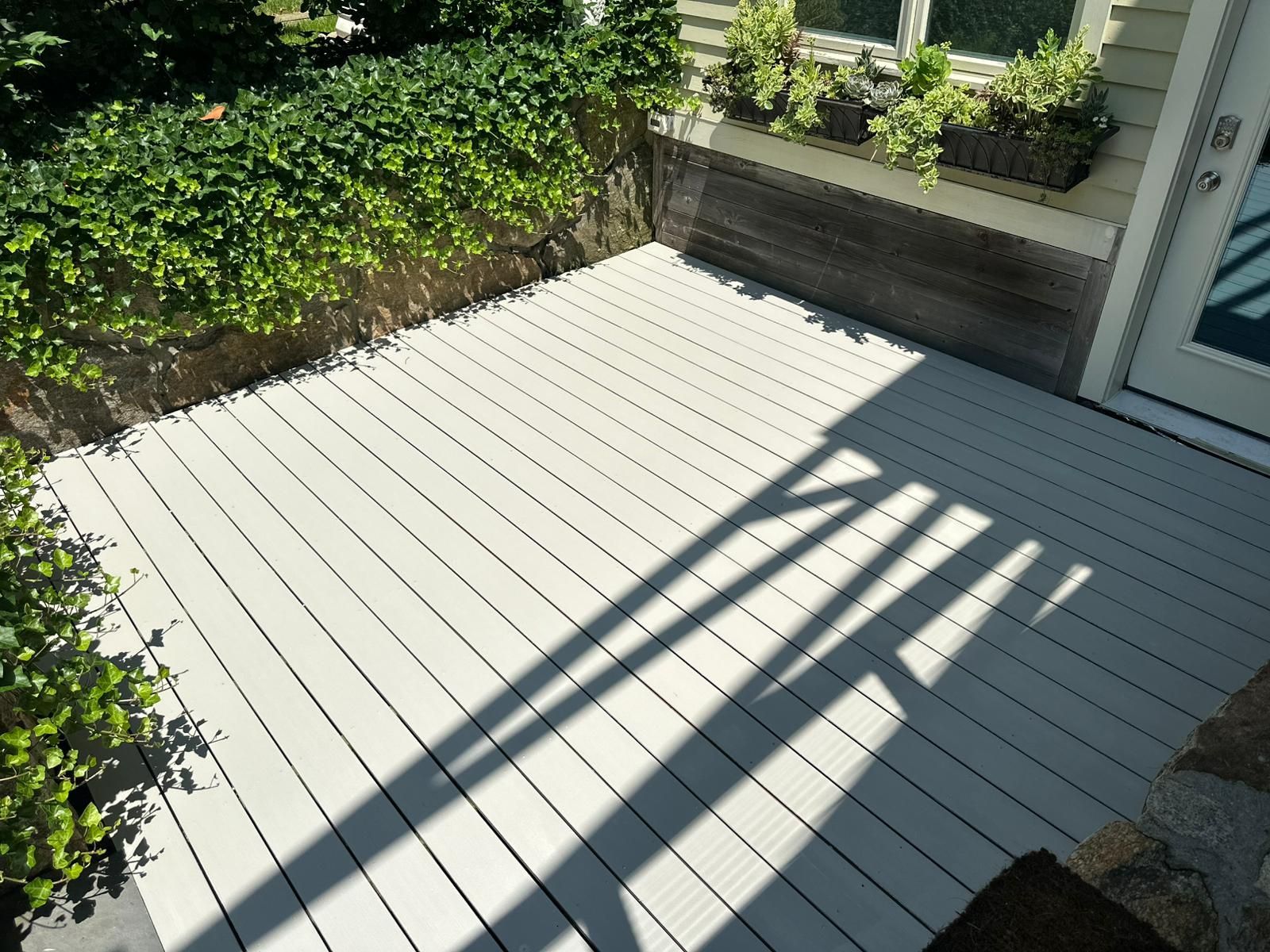 A small deck with light gray planks, bordered by green plants, next to a doorway.