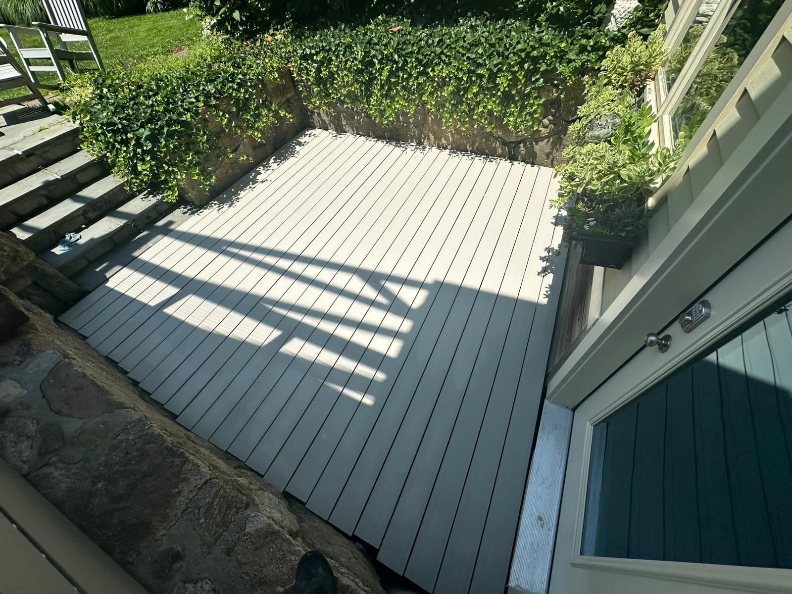 A deck with light-colored boards is surrounded by stone walls and greenery. Doorway on the right.