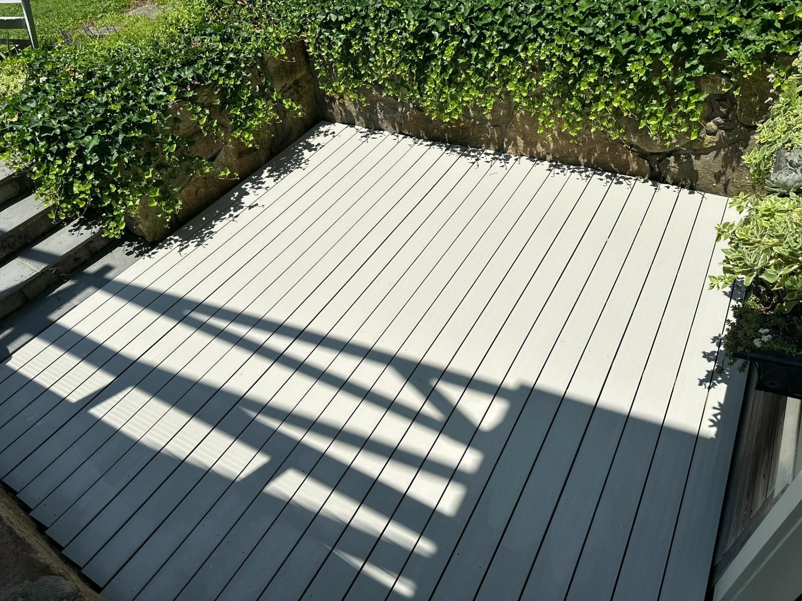 A light-colored wooden deck surrounded by a stone wall with green ivy. Sunlight creates shadows.