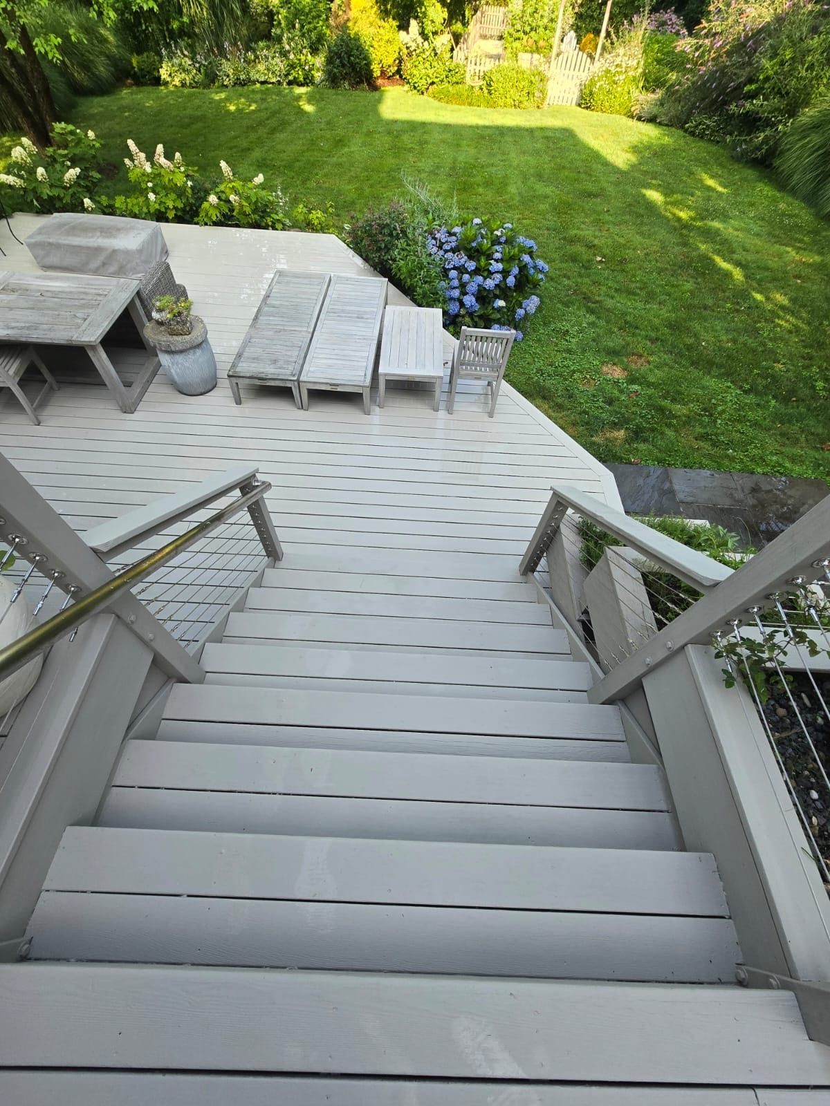 Wooden stairs leading down to a gray deck with lounge furniture and a green lawn with blooming blue flowers.