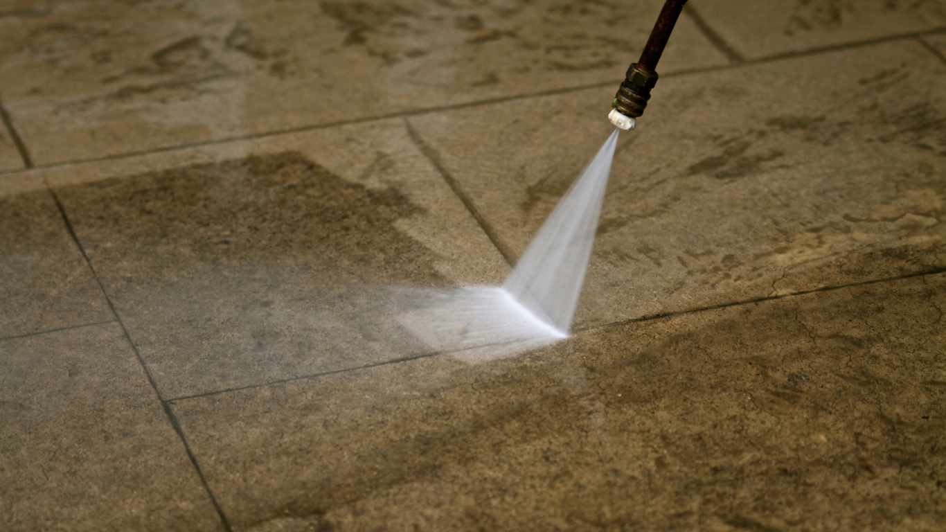 A person using a pressure washer to clean stone patio tiles. Water sprays from the nozzle, cleaning the surface.